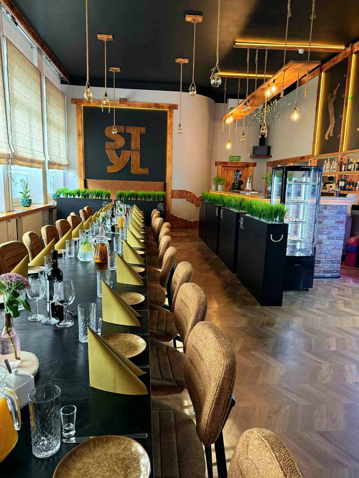 Sala restauracji: bar, wysokie krzesła przy oknach i logo STYL na ścianie